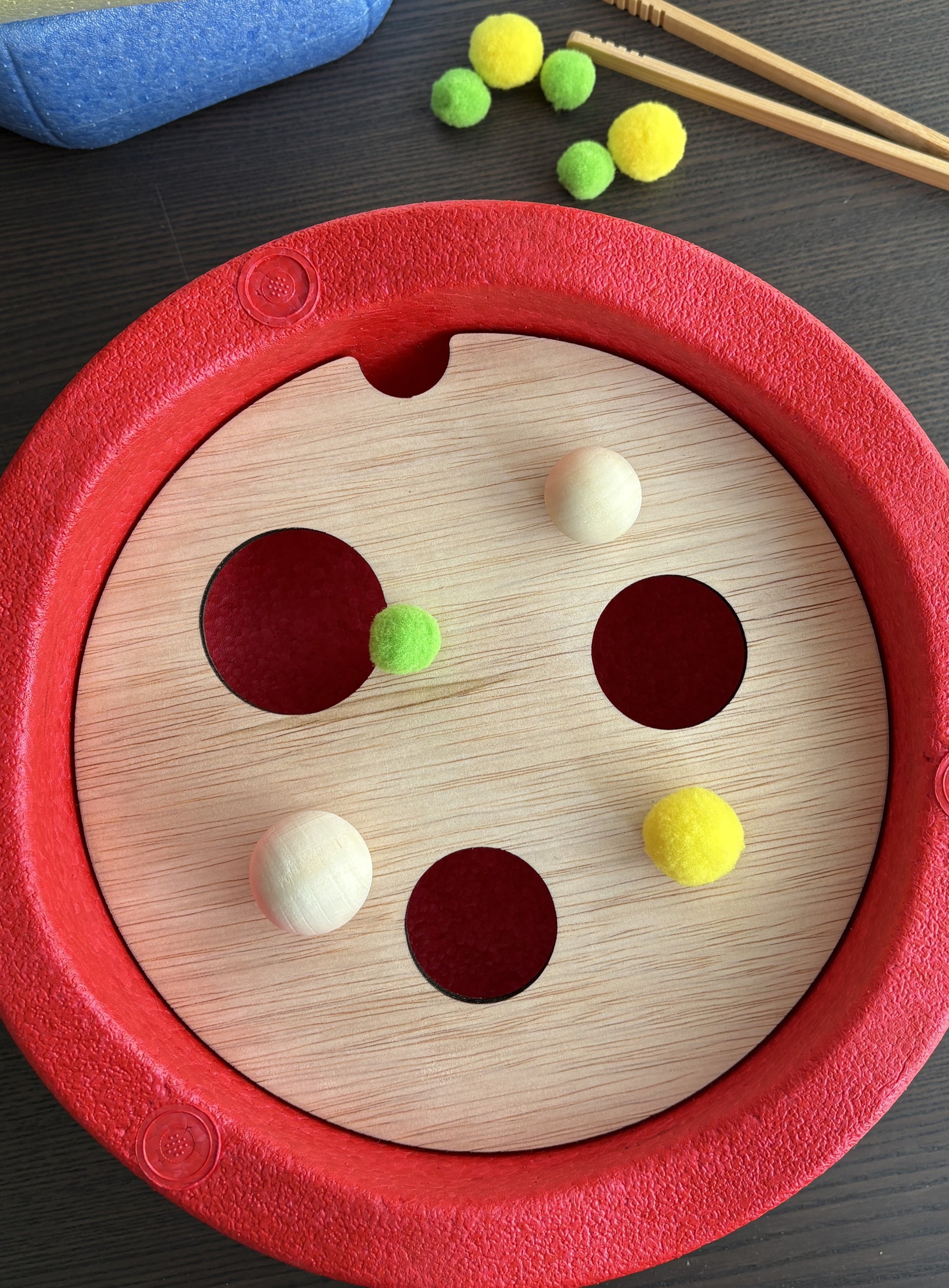 Houten plaat met drie gaten, in een rode ronde stapelsteen. Op de houten plaat liggen twee houten ballen een groene pompon en een grotere gele pompon. 