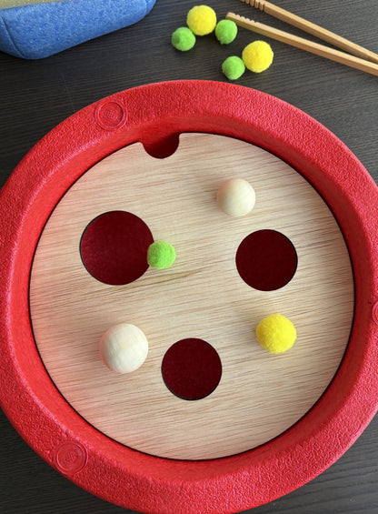 Houten plaat met drie gaten, in een rode ronde stapelsteen. Op de houten plaat liggen twee houten ballen een groene pompon en een grotere gele pompon. 